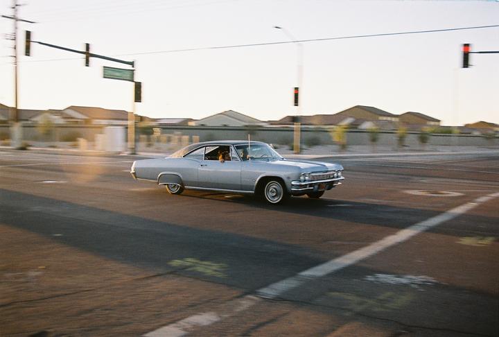 A silver vintage car with a dog in the passenger seat drives down a street at sunset.