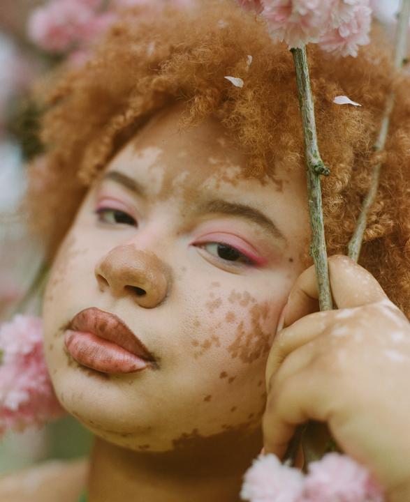 A woman with curly red hair and pink eye makeup holds a branch with pink blossoms.