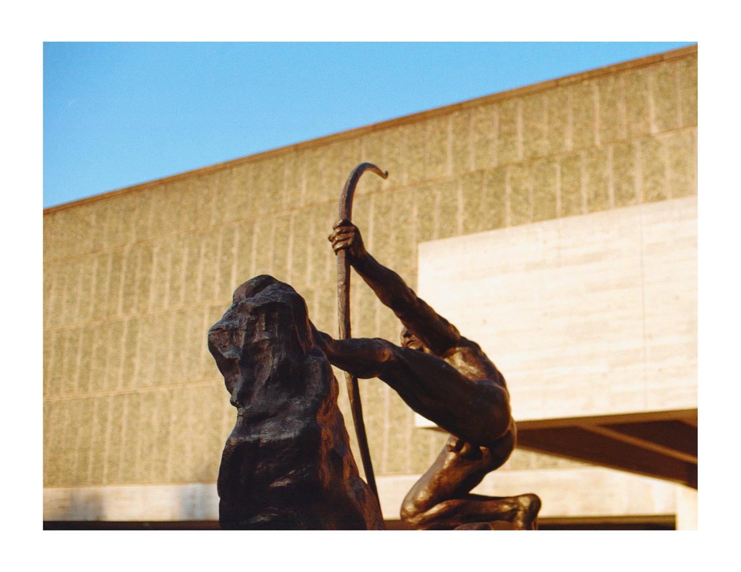 A bronze statue of a man with a bow is positioned in front of a building under a clear sky.