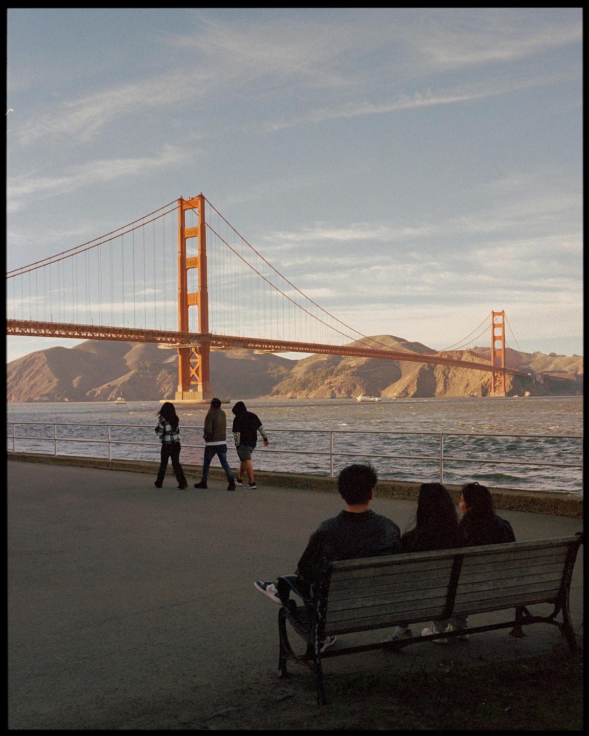 Three people sit on a bench looking at the Golden Gate Bridge while three other people walk along the water.