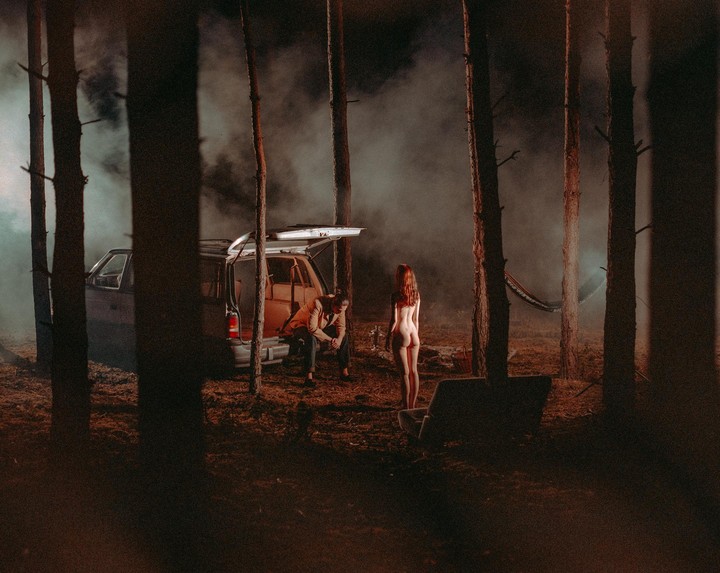 A nude woman stands in a foggy forest with a man sitting in the back of a car.