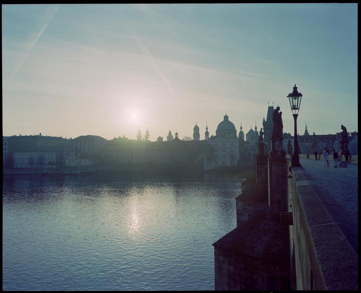 The sun rises over a city skyline with a river in the foreground and a bridge with statues and a lamppost on the right.