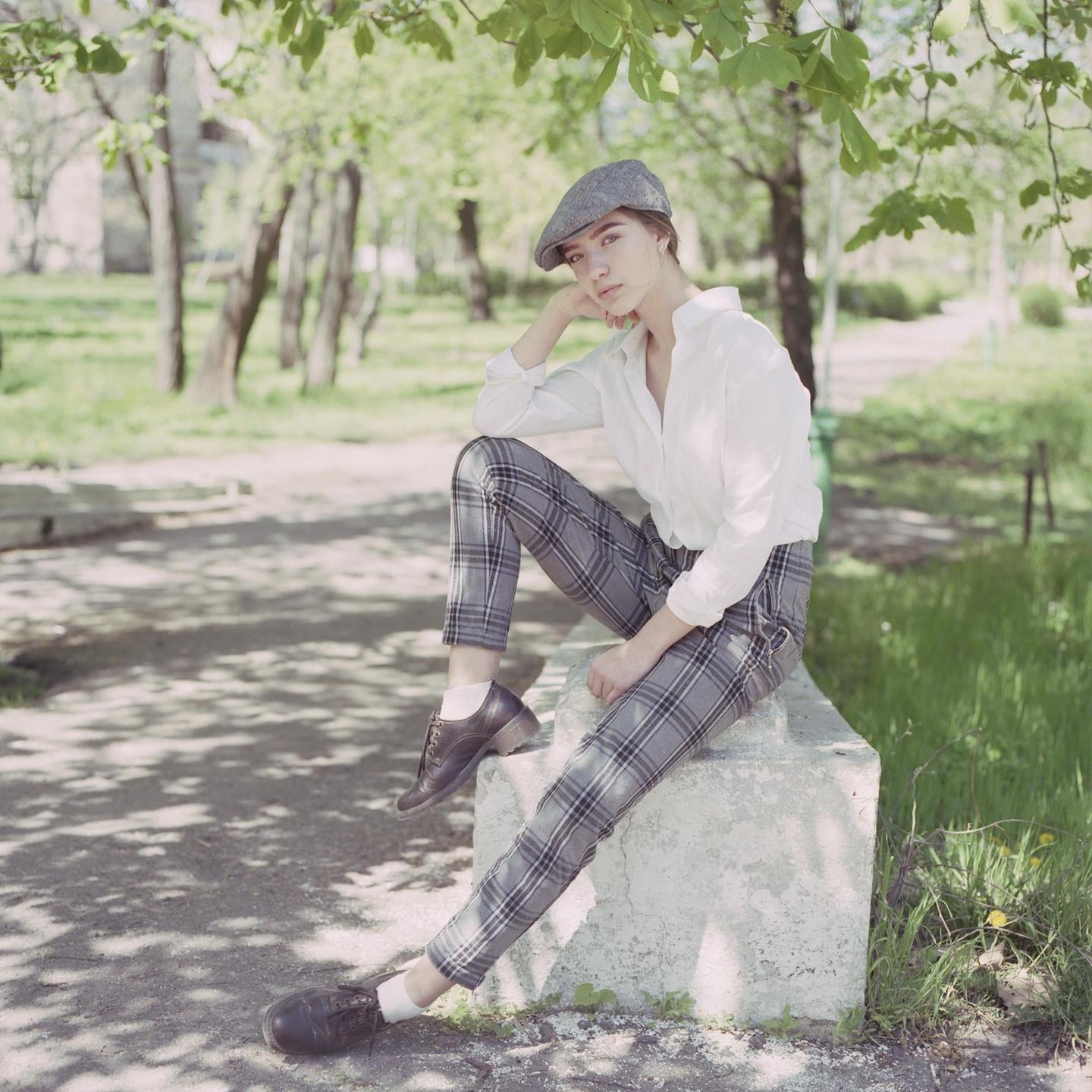 A woman wearing a flat cap and plaid trousers sits on a concrete block in a park.
