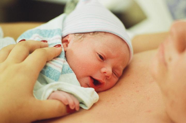 A newborn baby with a striped hat and blanket is held close to an adult's chest, with a hand gently touching the blanket.