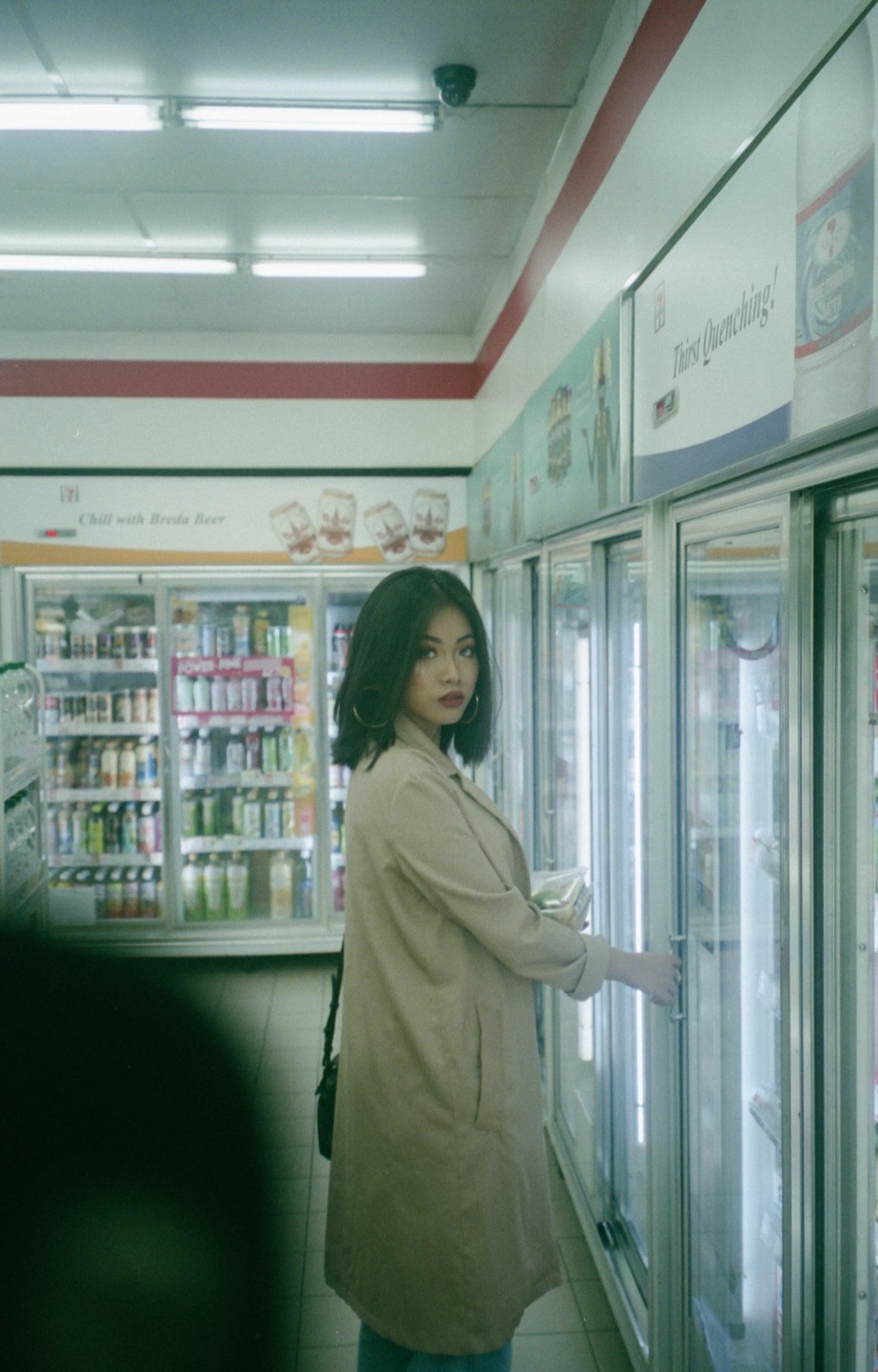 A woman in a trench coat stands in a convenience store aisle, looking towards the camera while holding a refrigerator door open.