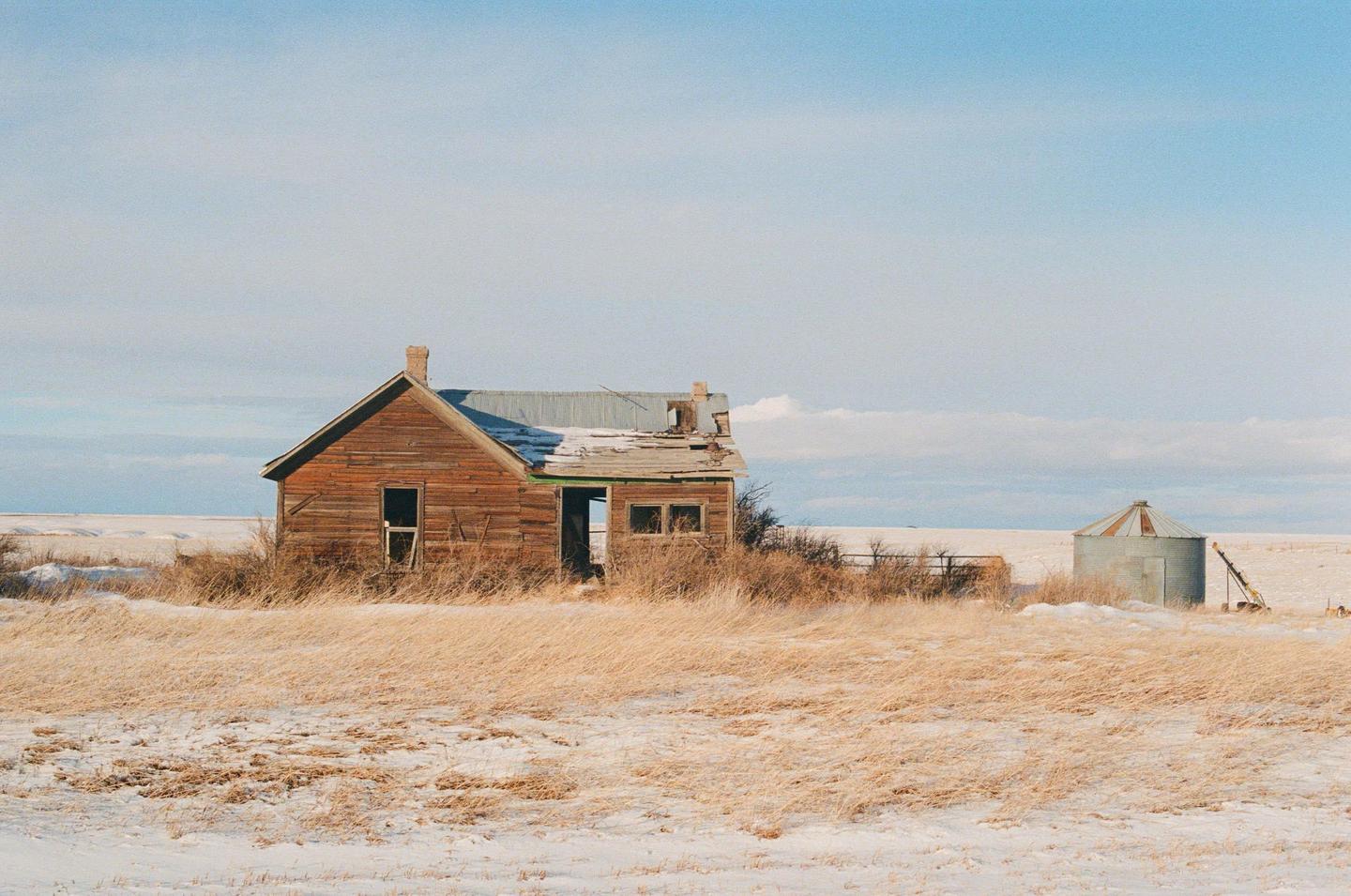 An abandoned wooden house and a small silo stand in a snowy field with dry grass under a pale blue sky.
