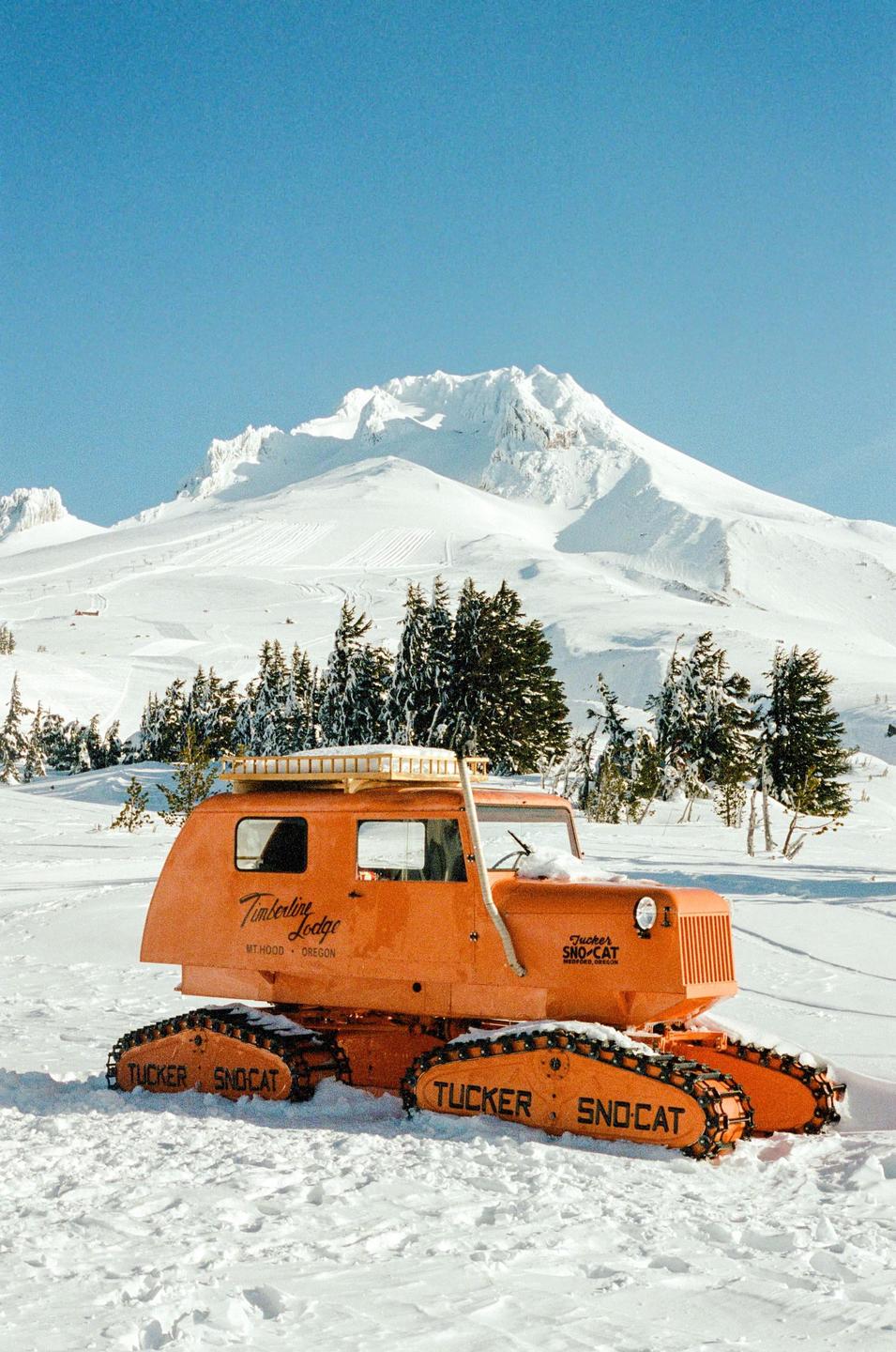 An orange snowcat is parked in the snow with a mountain and trees in the background.