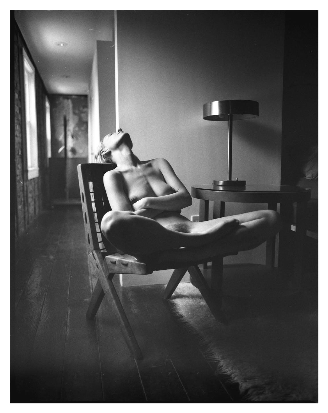 A nude woman sits in a wooden chair with her head tilted back and eyes closed.