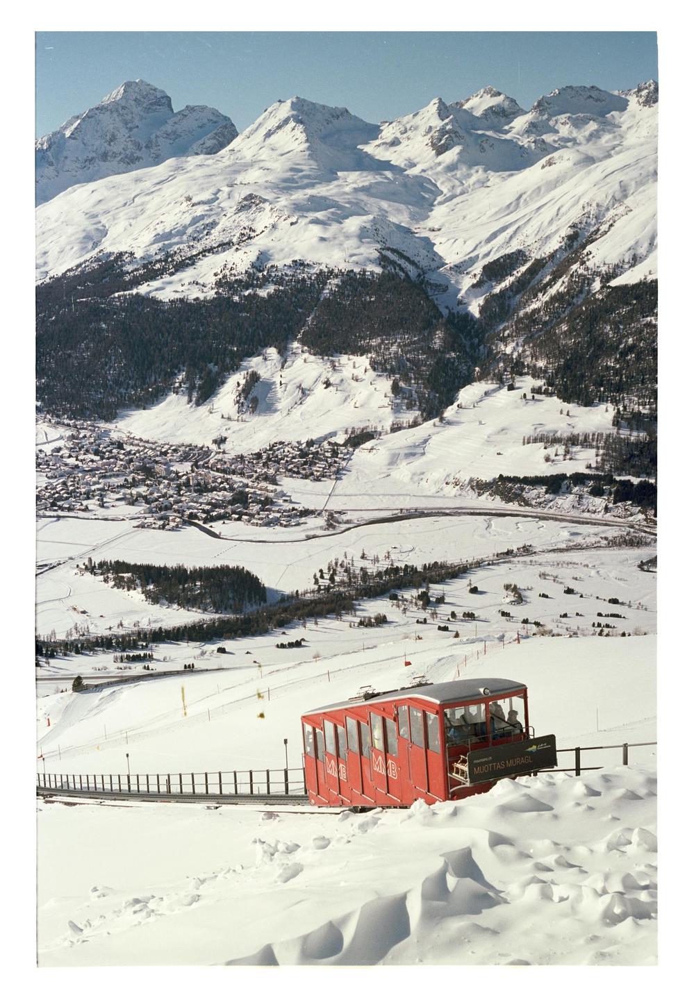 A red funicular car ascends a snowy mountain with a village and more mountains in the background.