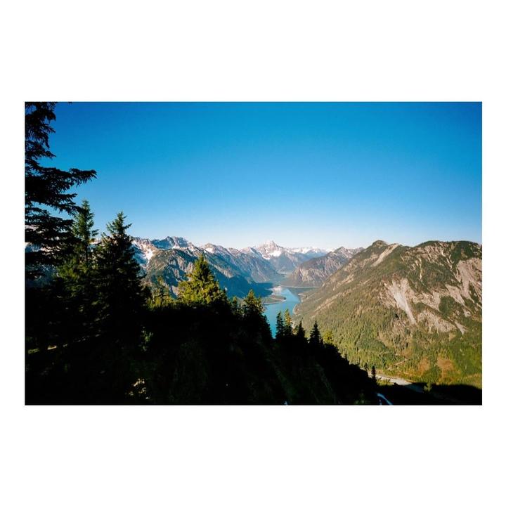 A winding river flows through a valley surrounded by mountains and evergreen trees under a clear blue sky.