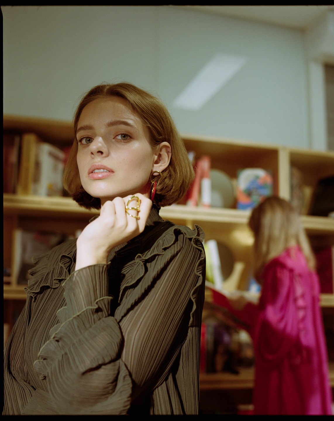 A woman with a gold ring and red earring poses in front of a bookshelf while another woman in pink looks at books in the background.