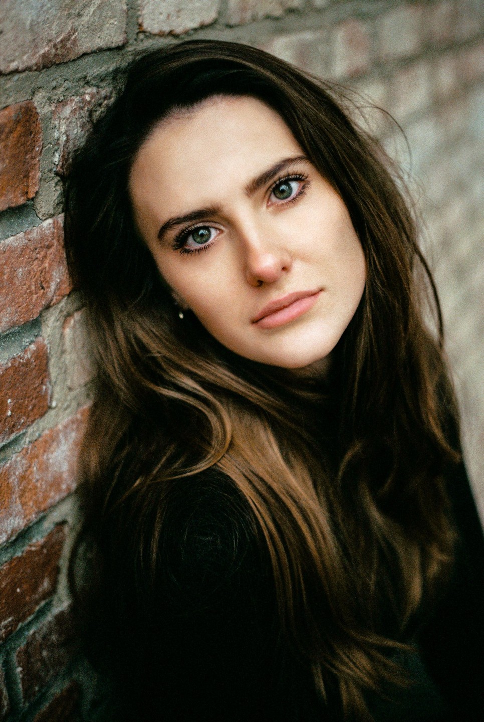A woman with long hair leans against a brick wall and looks at the viewer.
