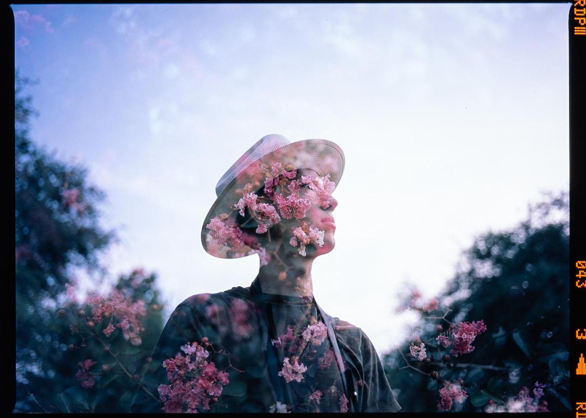 A man in a hat is double exposed with pink blossoms against a bright sky.