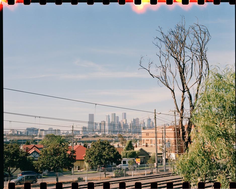 A city skyline is visible in the distance behind residential buildings, trees, and railroad tracks.