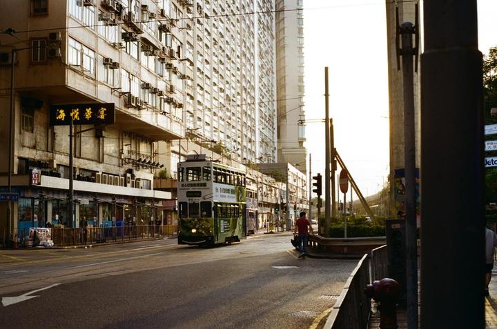 A green double-decker tram travels down a city street lined with tall buildings and a man walks on the sidewalk.