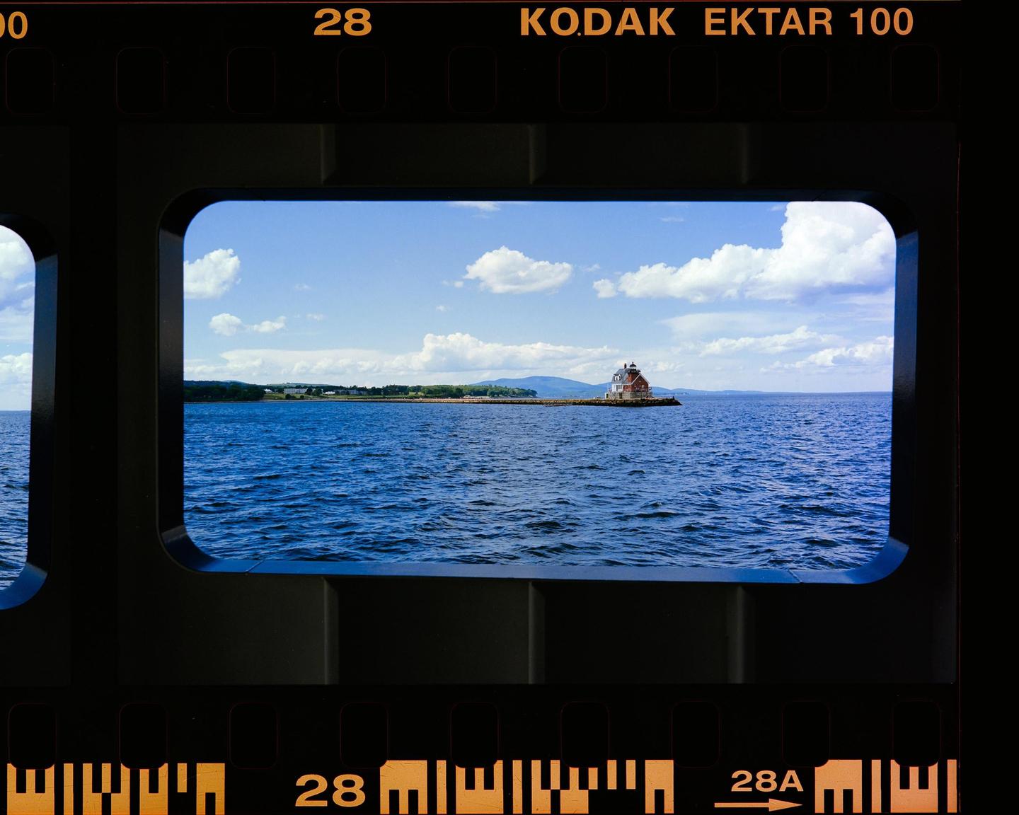 A small house sits on a rocky outcrop in the middle of a large body of water under a blue sky with white clouds.