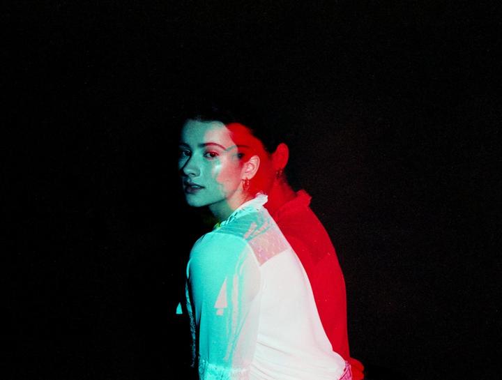 A woman in a white dress looks over her shoulder with red and blue light illuminating her.