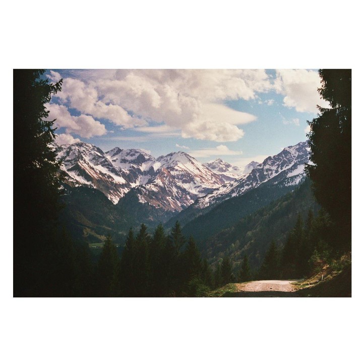 A mountain range with snow-capped peaks is framed by dark evergreen trees under a cloudy sky.