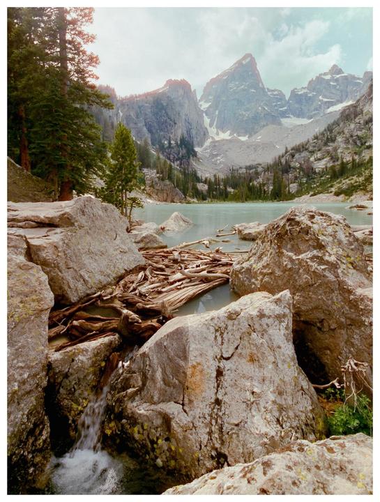 A mountain lake with driftwood and a small waterfall is surrounded by rocky peaks and pine trees.