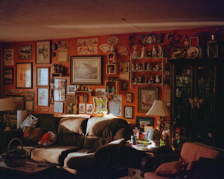 Sunlight illuminates a cluttered living room with a brown sofa, a display cabinet, and an orange wall covered in framed pictures and figurines.