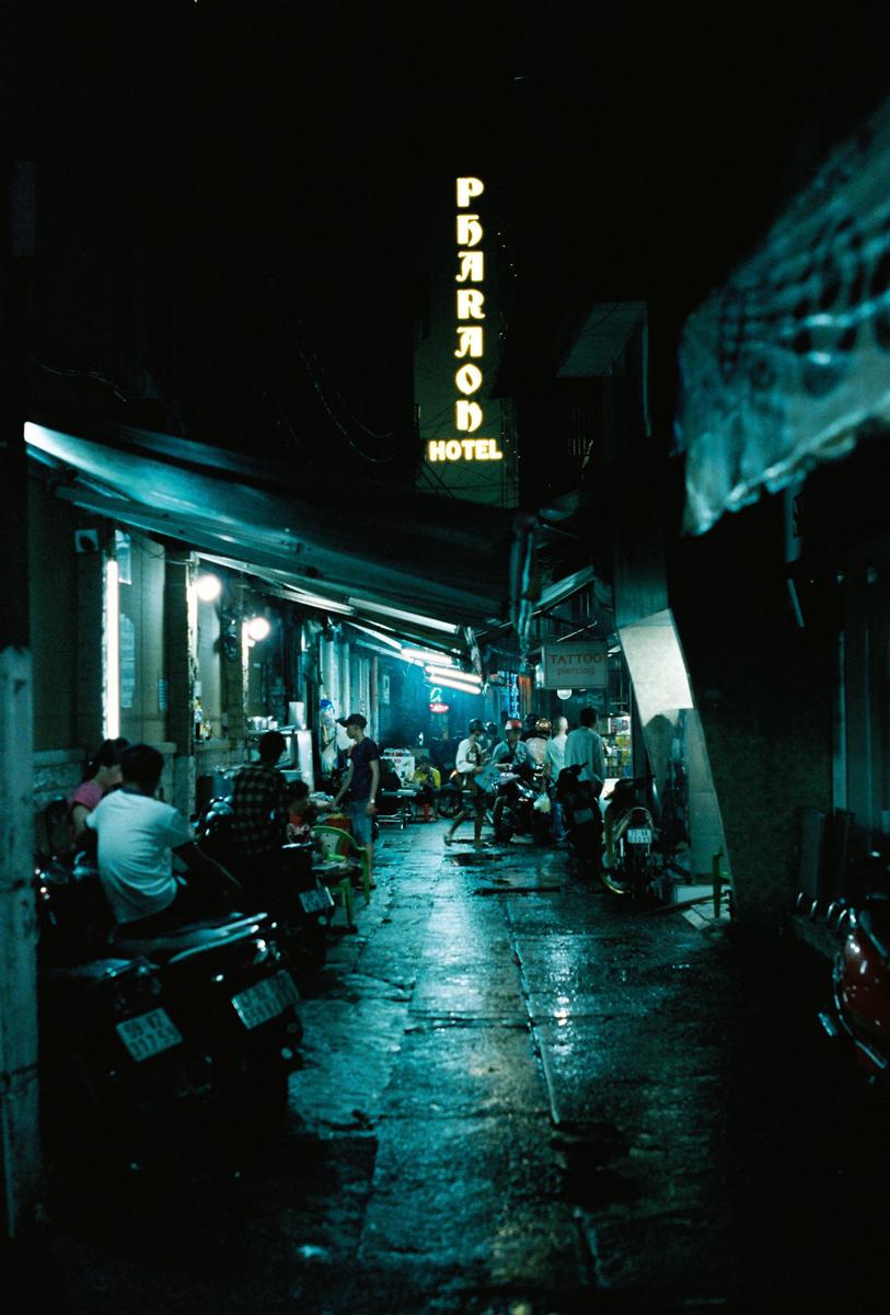 A neon sign for the Pharaon Hotel illuminates a wet street with people and motorbikes at night.
