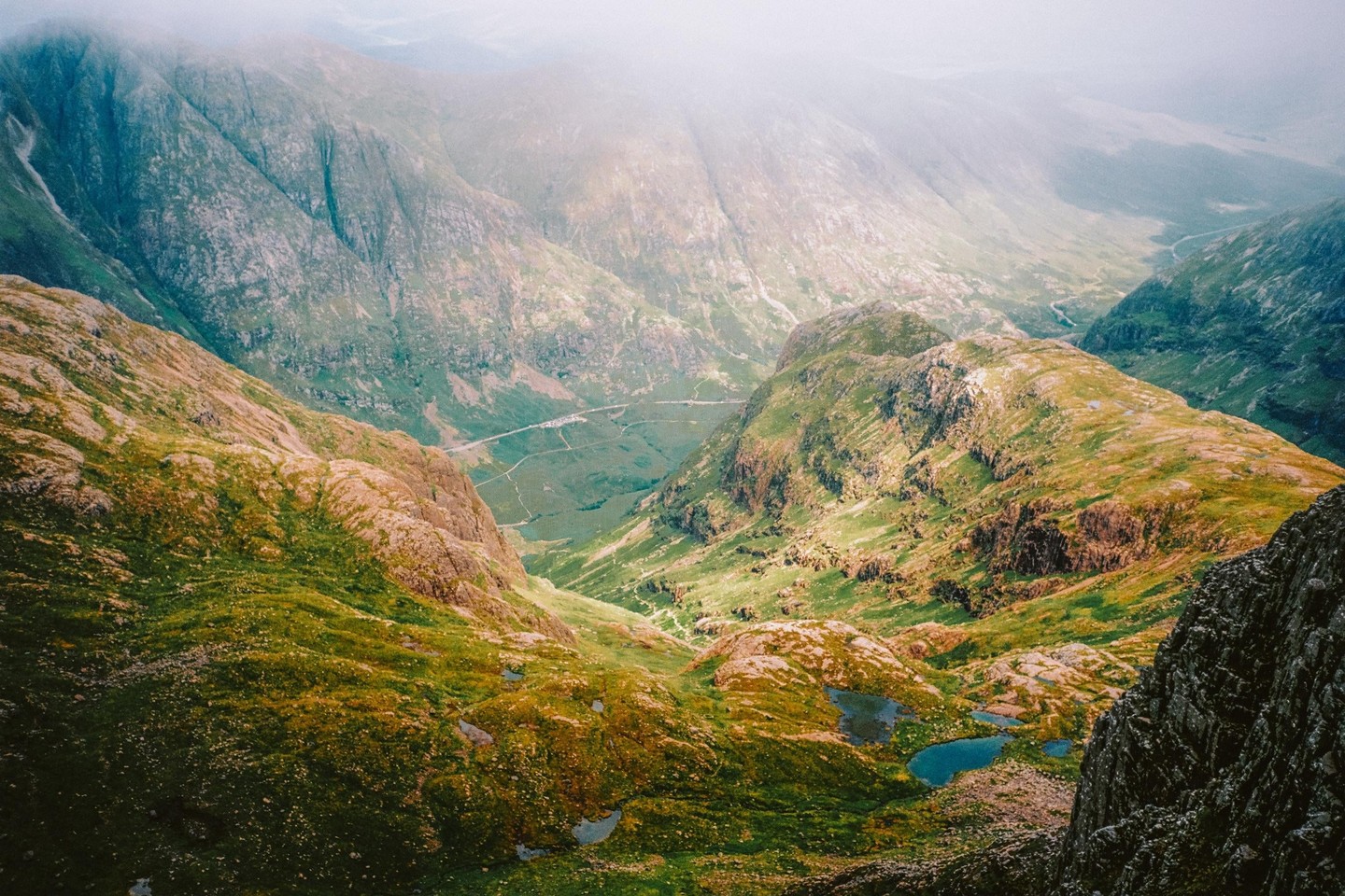 A hazy mountain landscape shows a valley with a road and small lakes.