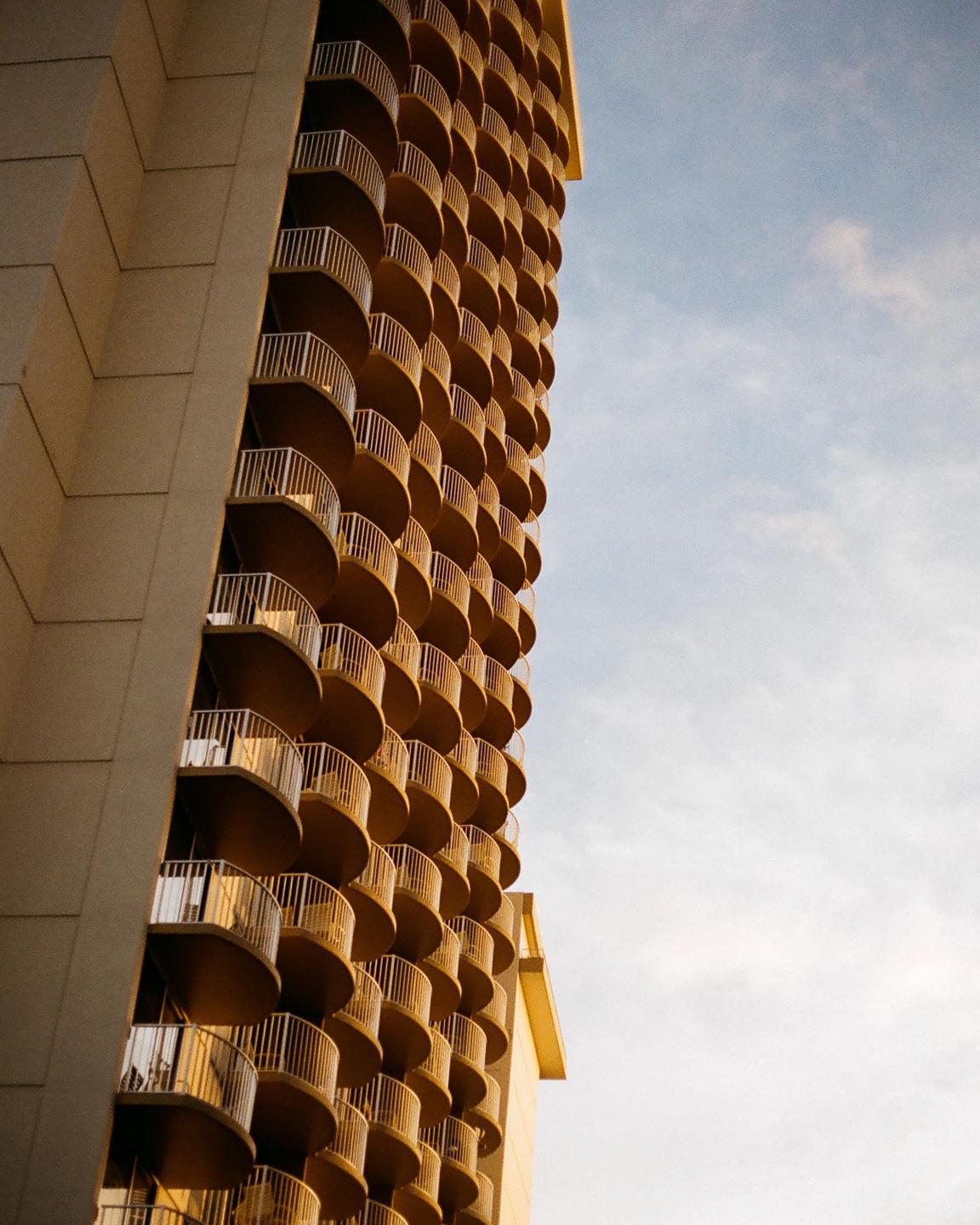 A tall building with many curved balconies is shown against a partly cloudy sky.