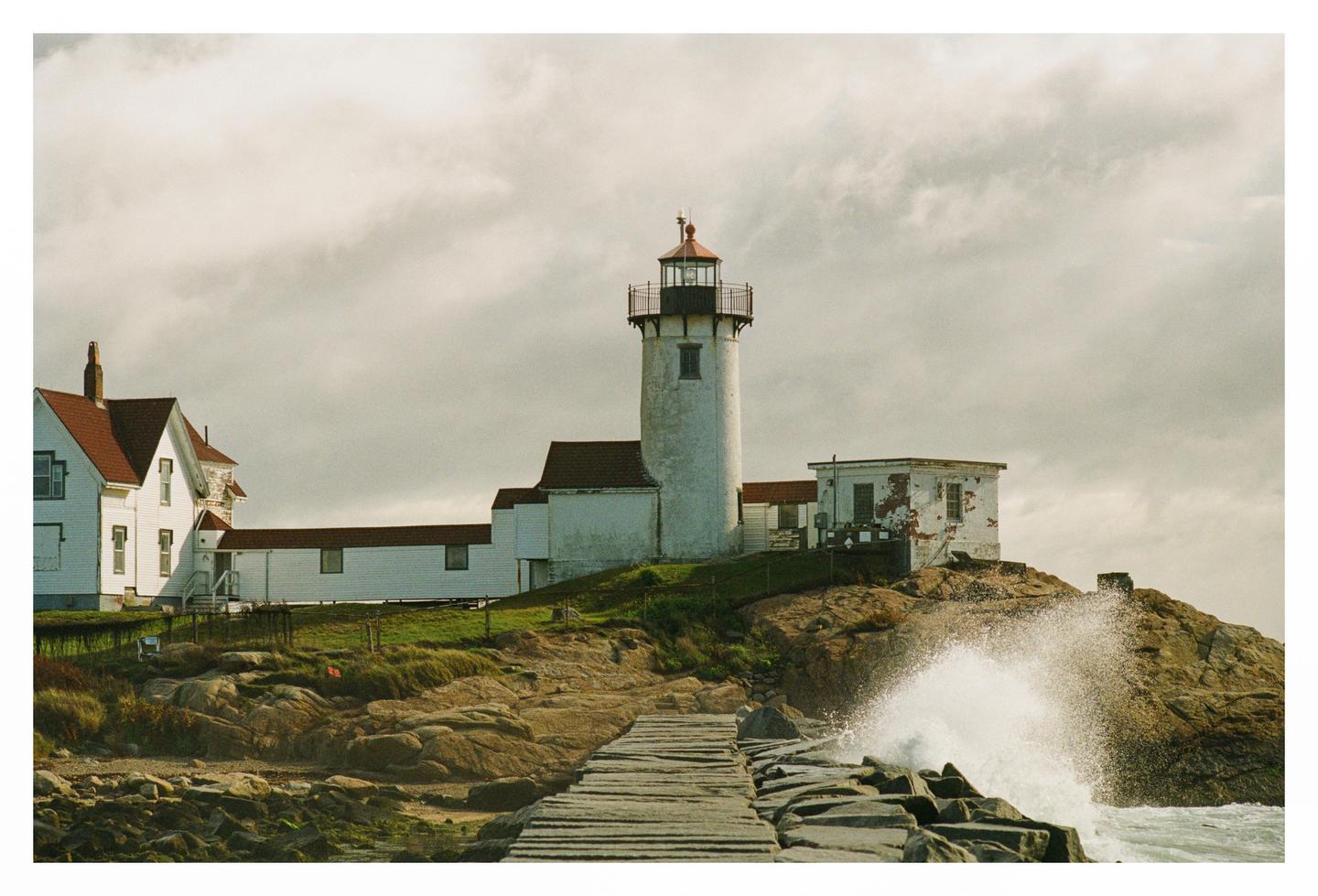 A white lighthouse and buildings stand on a rocky hill next to a wooden pier with a wave crashing against the rocks.