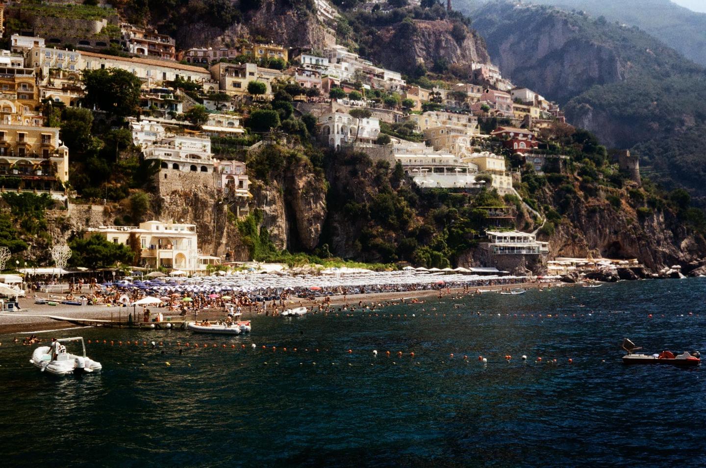 A hillside town with many buildings overlooks a crowded beach and the sea with boats.
