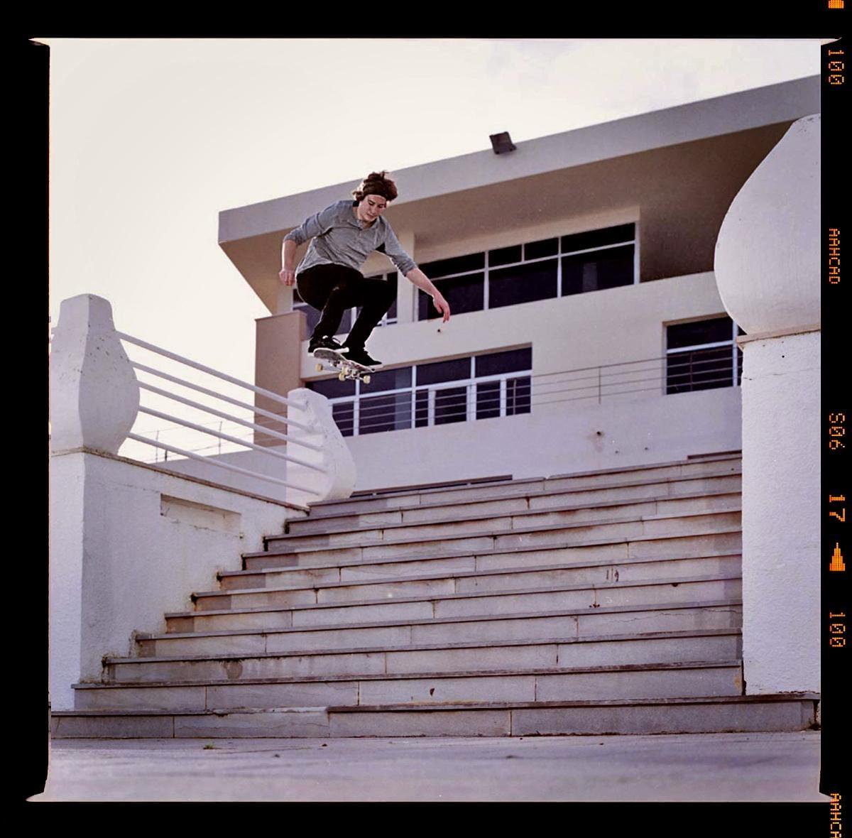 A man on a skateboard is airborne above a concrete staircase with a building in the background.