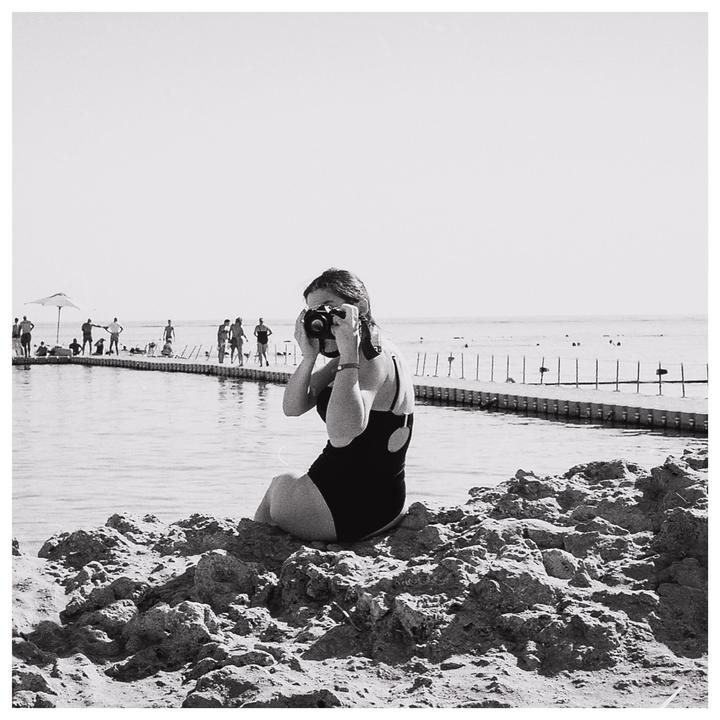 A woman in a swimsuit sits on rocks by the water and holds a camera to her face.