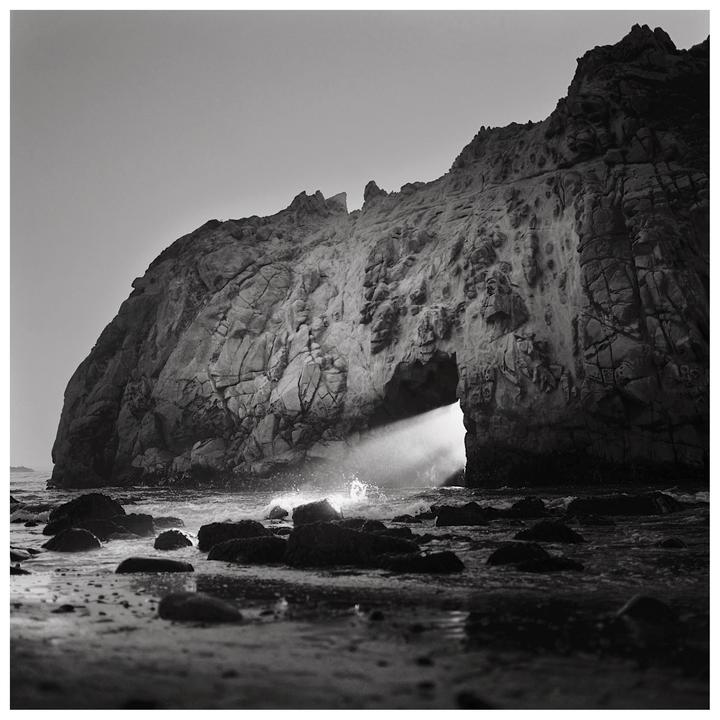 A beam of light shines through a keyhole arch in a large rock formation on a beach with waves crashing on the shore.