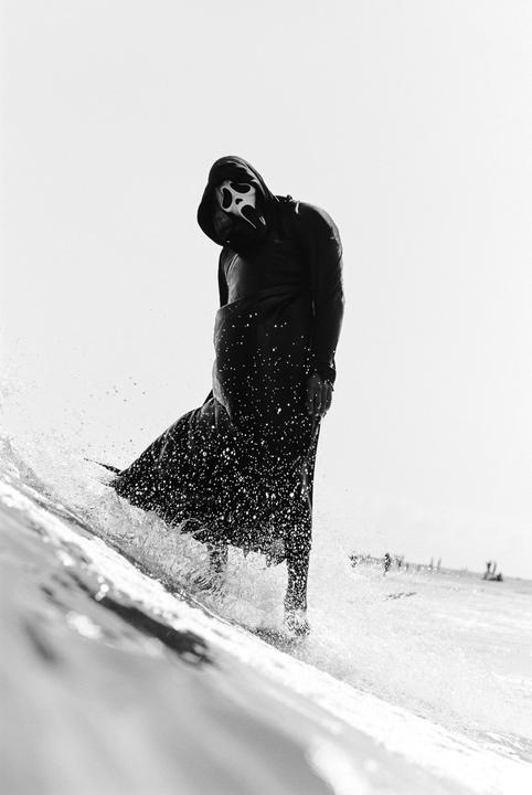 A person in a ghostface costume surfs on a body of water, creating a splash.