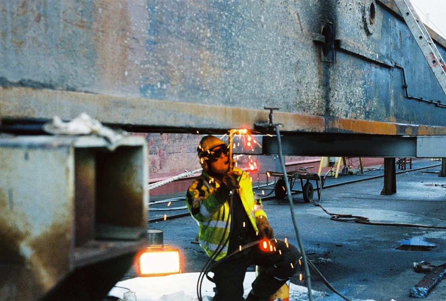 A man in a hard hat and safety vest welds a large metal structure, creating bright sparks.