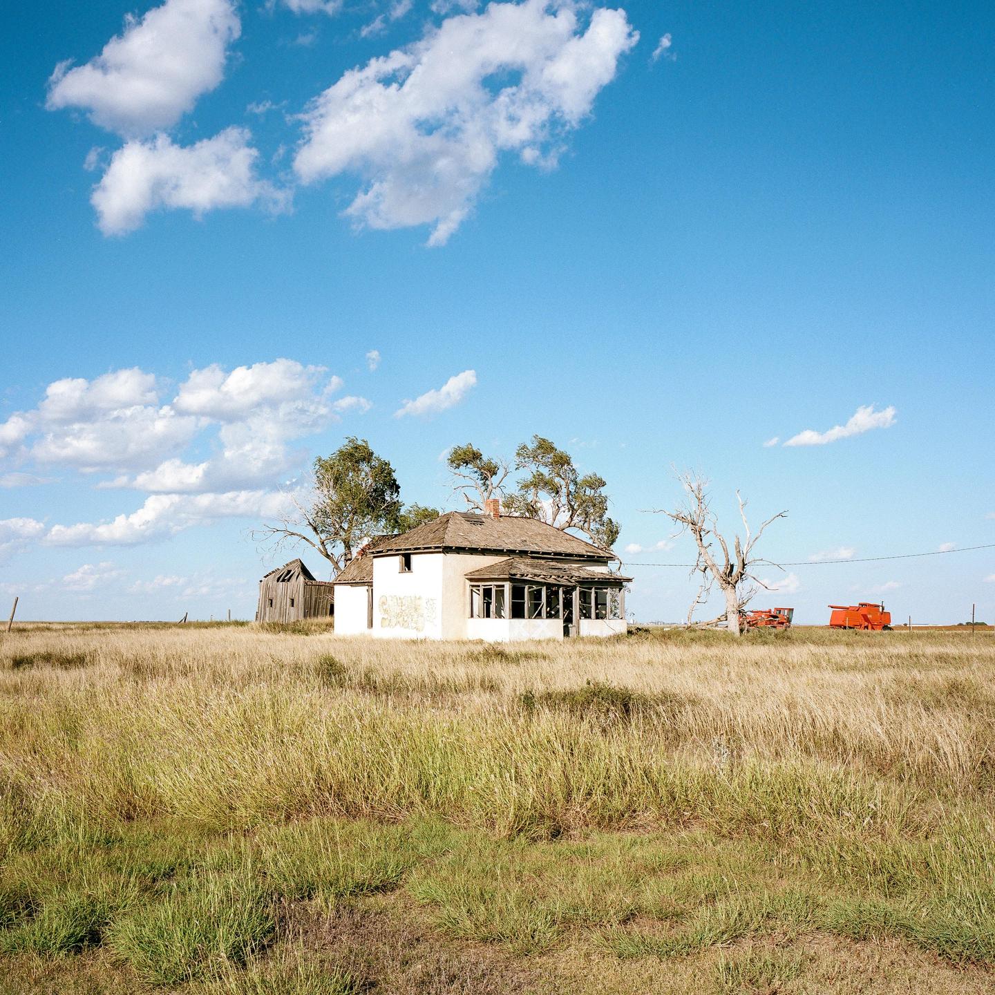 An abandoned house stands in a field of tall grass under a blue sky with scattered clouds.