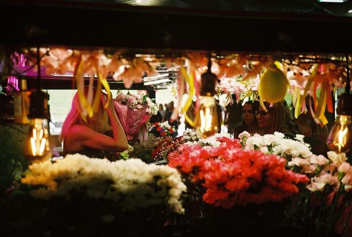 A woman with blonde hair is visible behind a display of flowers and hanging light bulbs.