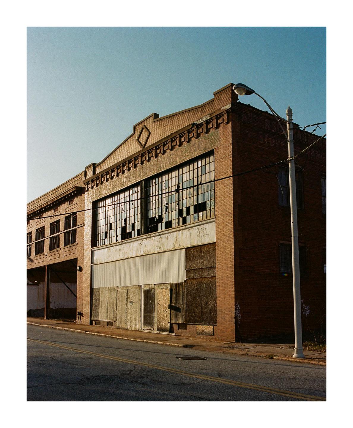 An abandoned brick building with broken windows and boarded-up lower levels stands beside a street with a lamppost.