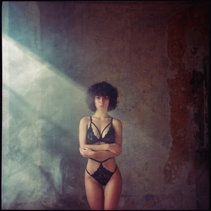 A woman with curly hair stands in black lingerie with her arms crossed in a hazy room with a beam of light.