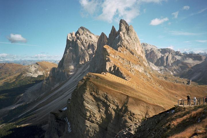 A panoramic view of the Seceda mountain range in the Dolomites shows jagged peaks and a valley with a few people on a viewing platform.