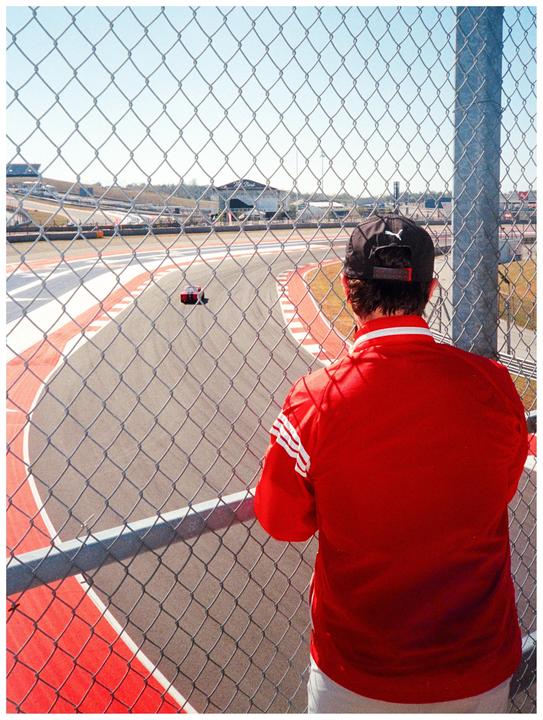 A man in a red shirt and black cap watches a race car on a track through a chain link fence.