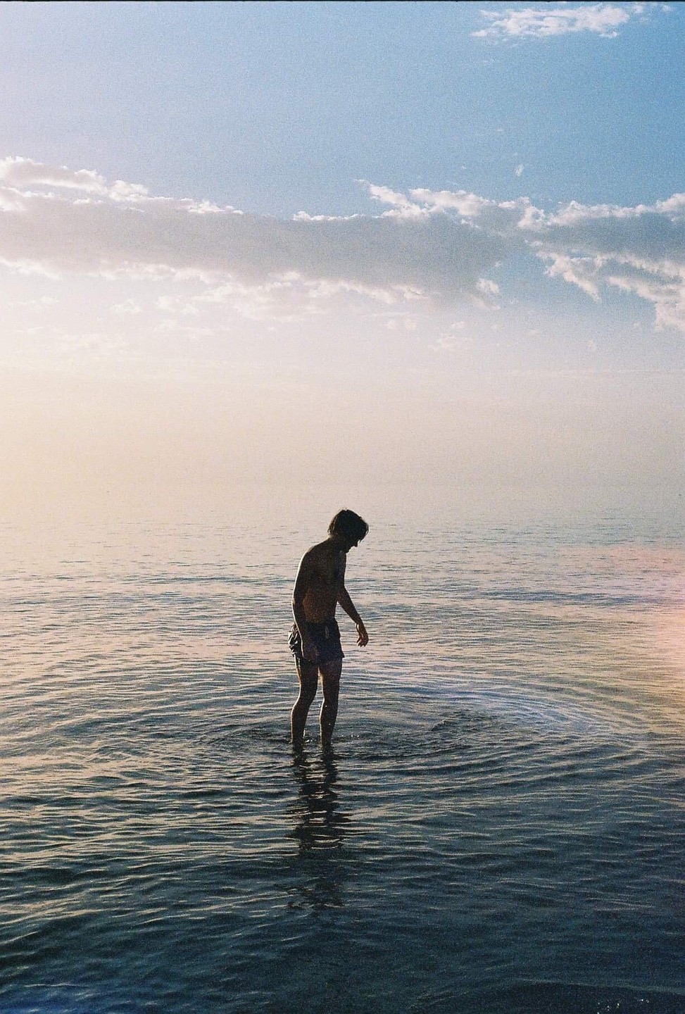A man stands in calm water under a hazy sky with scattered clouds.