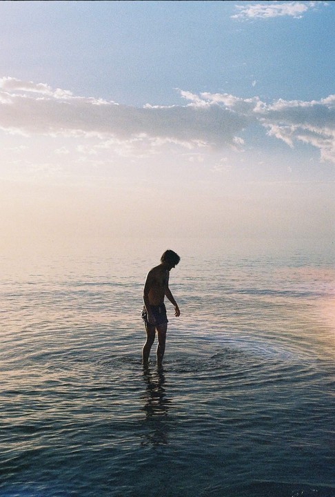 A man stands in calm water under a hazy sky with scattered clouds.