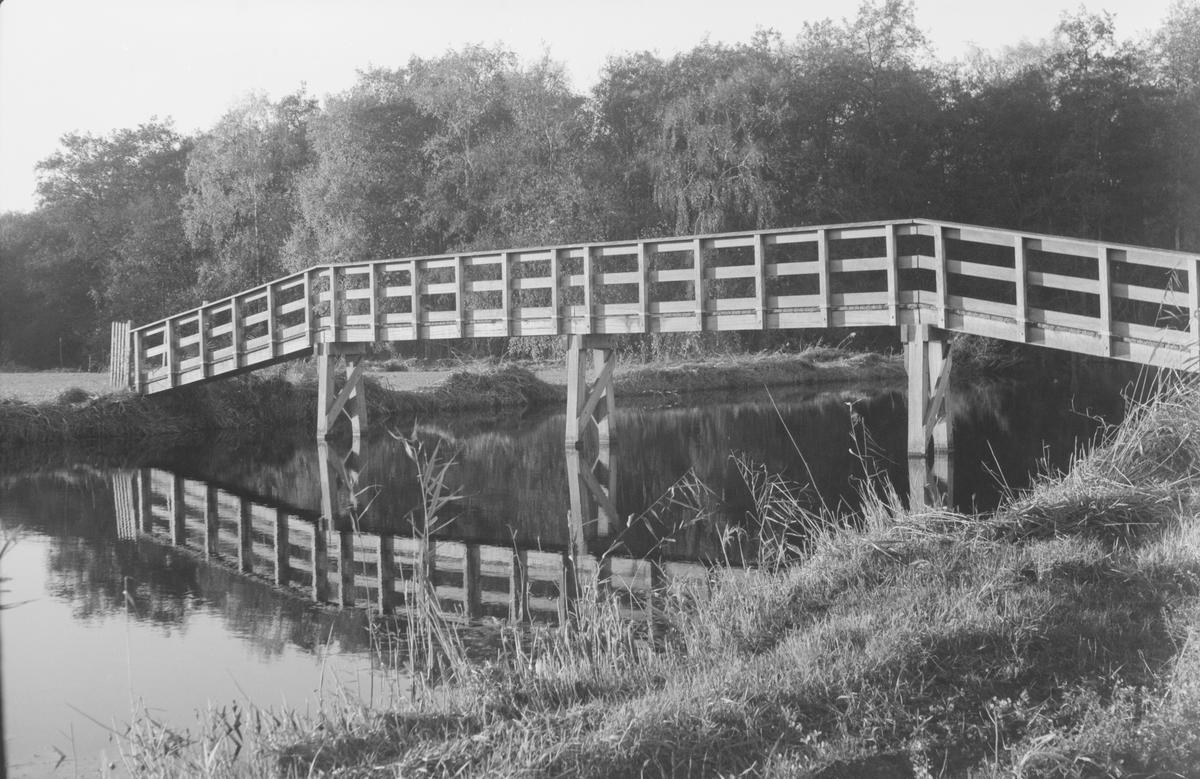 A wooden footbridge crosses a narrow body of water, reflecting in the still surface.