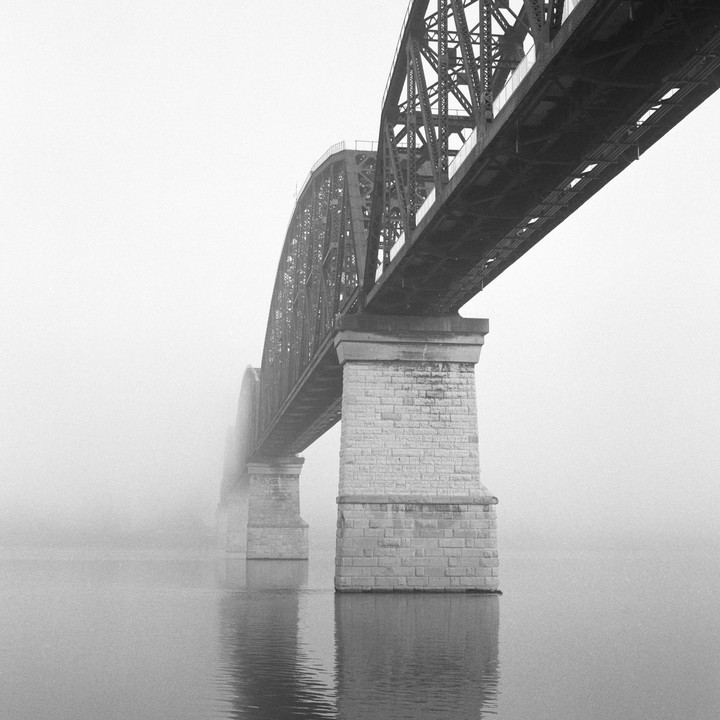 A large metal bridge with stone piers extends over a body of water shrouded in fog.