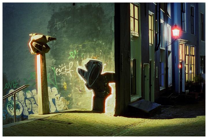 A street scene at night shows a stone sculpture, graffiti, and illuminated windows and a street lamp.