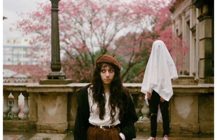 A woman in a beret stands next to a person dressed as a ghost on a stone balcony with pink blossoms in the background.