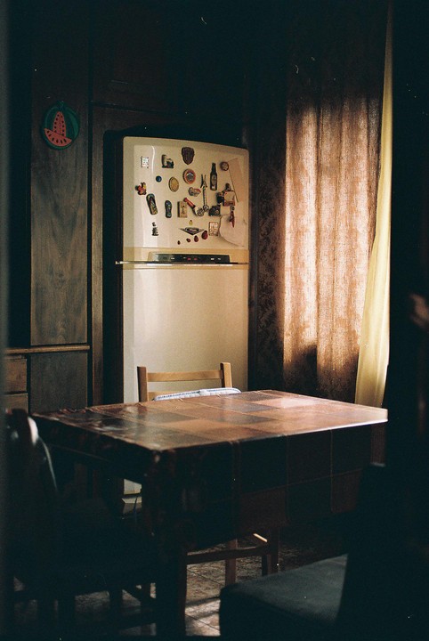 A refrigerator covered in magnets stands next to a window with curtains, with a table and chair in the foreground.