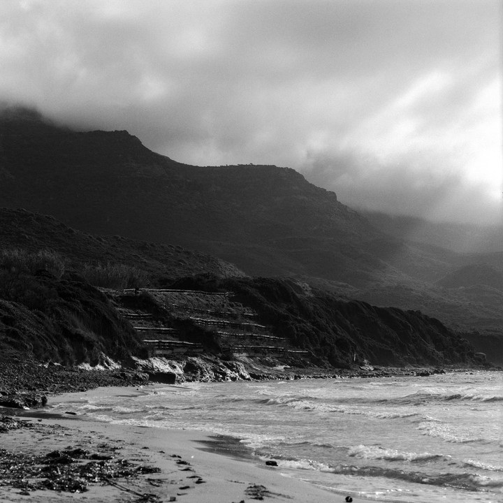 A black and white landscape shows a beach with waves crashing on the shore, with a large mountain covered in vegetation in the background.
