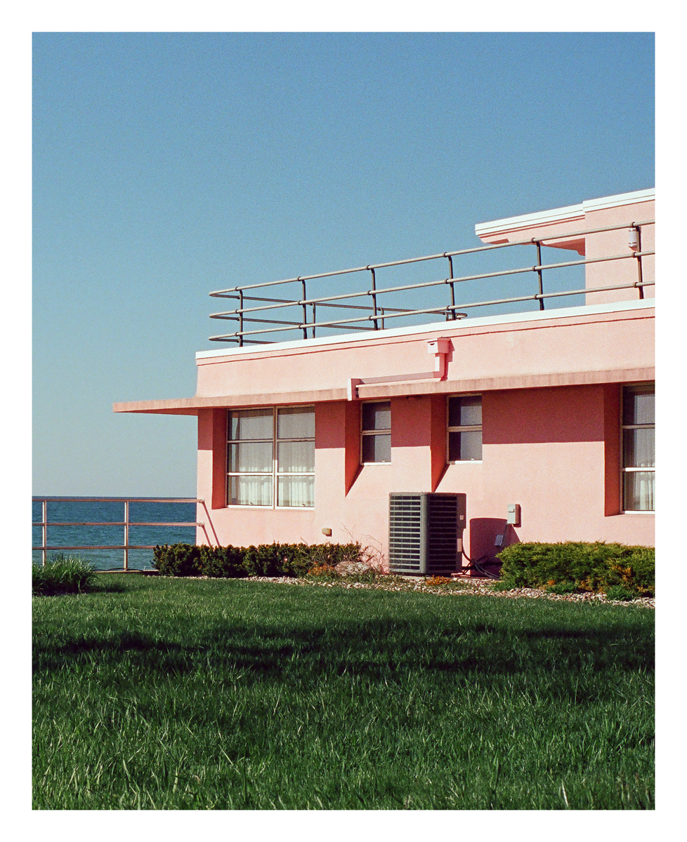 A pink building with a flat roof and railing stands next to a body of water, with green grass in the foreground.