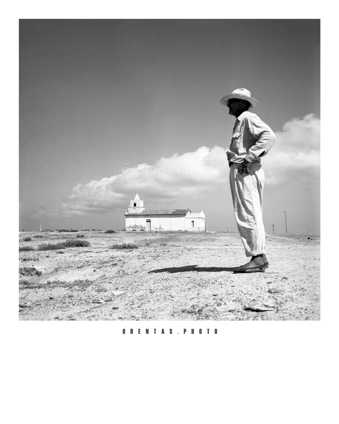 A man in a hat stands in a barren landscape with a small church in the background under a cloudy sky.
